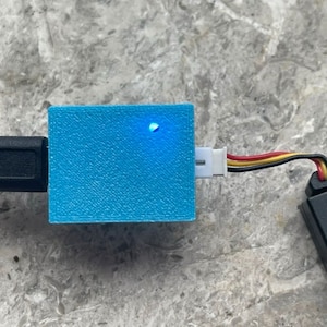 Könnte beinhalten: Eine kleine, hellblaue, 3D-gedruckte Box mit einer blauen LED-Leuchte oben. Die Box ist mit einem schwarzen USB-Kabel und einem schwarzen und gelben Kabel verbunden. Das Kabel ist mit einem schwarzen Akku verbunden.