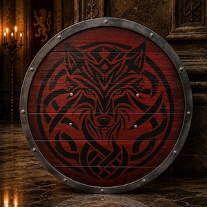 Pode incluir: Um escudo circular de madeira com acabamento tingido em vermelho escuro e um gráfico proeminente de cabeça de lobo preta com padrões de nós intrincados. O escudo é contornado por uma borda de prata metálica fixada com rebites espaçados.