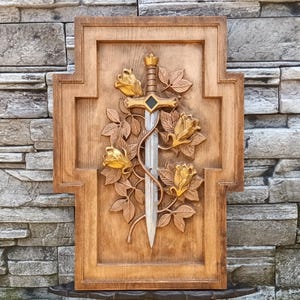 Op de afbeelding: Houten wandkunst met een gebeeldhouwd zwaard omwonden met rozen. Het zwaard heeft een gouden handvat en een zilveren lemmet, met drie gouden rozen en bladeren. Het kunstwerk staat tegen een getextureerde, bruine houten achtergrond.