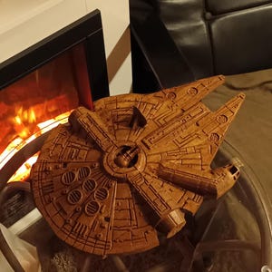 Può includere: Un modello in legno dettagliato della navicella spaziale Millennium Falcon, con intricati dettagli intagliati. Il modello marrone è posto su un tavolo con piano in vetro. La navicella è un'icona riconoscibile del franchise di Star Wars.