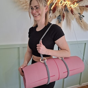 Könnte beinhalten: Eine gerollte, rosa Yogamatte mit grauen Gurten und Holzringen. Die Matte wird von einem geflochtenen grauen Gurt getragen. Im Hintergrund ein Neon-Schild mit der Aufschrift "Home" und ein floraler Wandbehang.