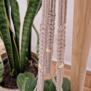 Könnte beinhalten: Ein beigefarbener Makramee-Pflanzenhänger mit Holzperlen. Der Hänger ist aus dickem Baumwollseil gefertigt und hat ein geknotetes Design.