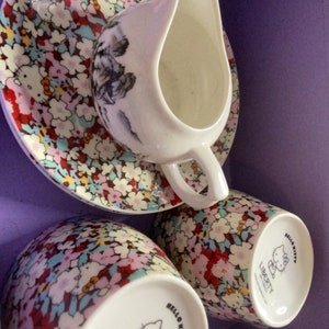 May include: A set of three Hello Kitty teacups and saucers with a floral pattern. The cups and saucers are white with a pink, blue, and green floral pattern. The cups have a Hello Kitty design on the bottom with the text "Hello Kitty" and "Liberty of London" printed on the bottom.
