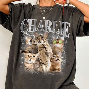 May include: Dark gray t-shirt featuring the name "CHARLIE" in a silver, outlined font above a graphic of five cats with lightning bolts. The cats have various expressions, with some looking up and others with wide eyes.