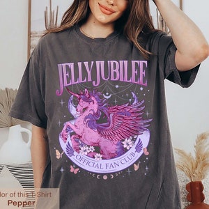 May include: A dark gray t-shirt with a graphic of a pink winged unicorn with a purple horn. The graphic is surrounded by stars, moons, and butterflies. The text "JELLY JUBILEE OFFICIAL FAN CLUB" is printed on the shirt.