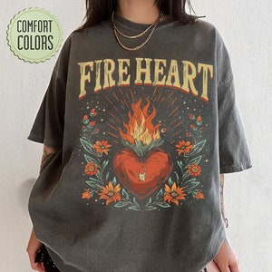 Peut inclure: Un t-shirt gris foncé avec le texte "FIRE HEART" en lettres jaunes et orange. Un cœur rouge avec des flammes est entouré de fleurs vertes et orange.