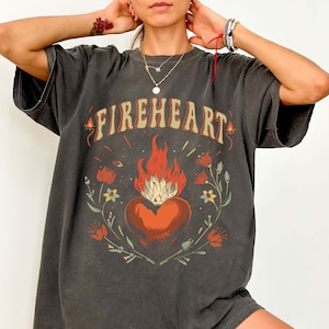 Puede incluir: Una camiseta gris antracita de gran tama&ntilde;o con un estampado gr&aacute;fico de un coraz&oacute;n en llamas rodeado de flores. El texto "FIREHEART" est&aacute; impreso sobre el coraz&oacute;n.