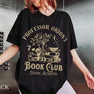 May include: Black t-shirt with a gold graphic design featuring a skull, flowers, and potion bottles. The text reads "Professor Orion's Book Club Zodiac Academy".