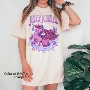 May include: A cream colored t-shirt with a graphic of a pink winged unicorn with a purple mane and tail. The graphic also includes the text "Jelly Jubilee Official Fan Club" in purple lettering.