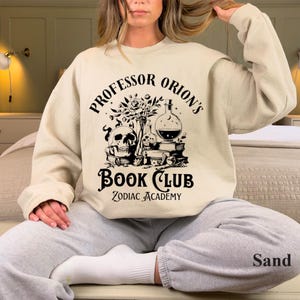 May include: A beige sweatshirt with a black graphic print that says "Professor Orion's Book Club Zodiac Academy". The graphic features a skull, flowers, a potion bottle, and books.