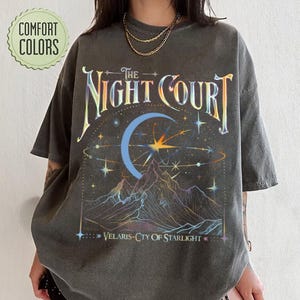 Op de afbeelding: Een donkergrijs T-shirt met een grafische print van een halve maan, sterren en bergen. De tekst "The Night Court" is in regenboogkleuren boven de print gedrukt. De tekst "Velaris - City of Starlight" is onder de print gedrukt.