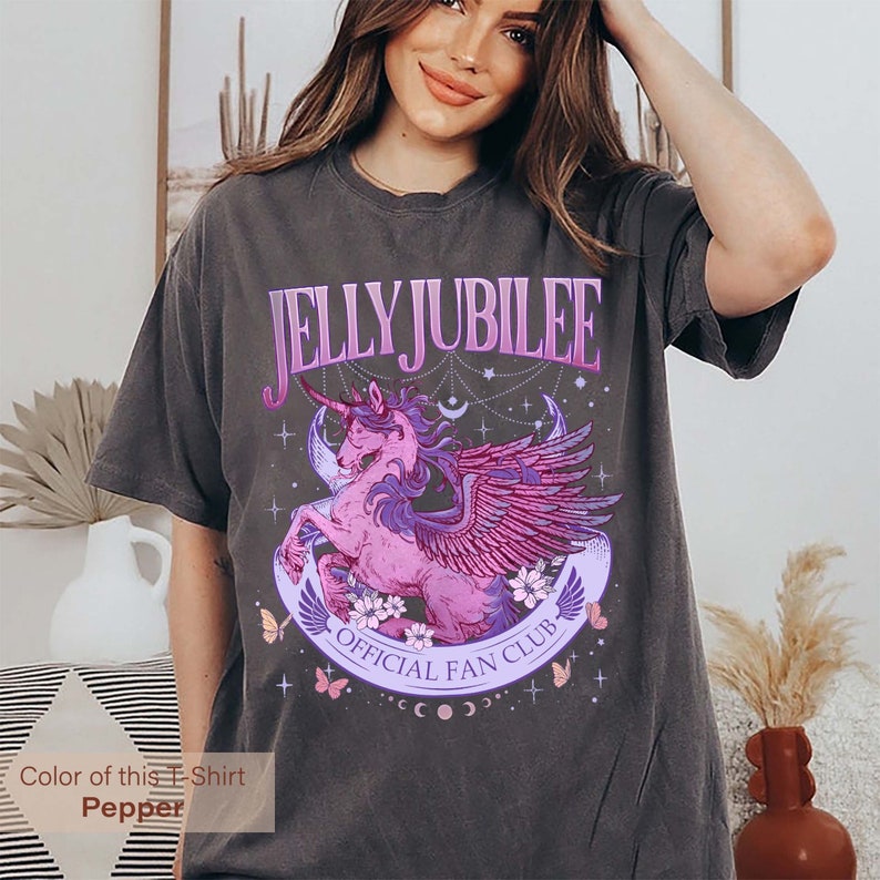 May include: A dark gray t-shirt with a graphic of a pink winged unicorn with a purple horn. The graphic is surrounded by stars, moons, and butterflies. The text "JELLY JUBILEE OFFICIAL FAN CLUB" is printed on the shirt.