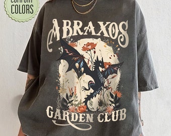 May include: A black t-shirt with a graphic print of a black dragon with wings, surrounded by flowers. The text "ABRAXOS GARDEN CLUB" is printed below the dragon.
