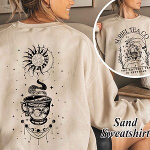 May include: A sand-colored sweatshirt with a black graphic of a teacup, moon, and sun. The text "Suriel Tea Co. The Hottest Tea in Prythian" is printed on the front of the sweatshirt.