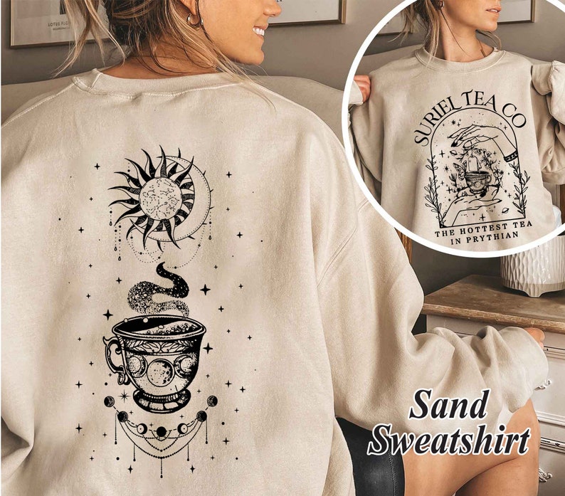 May include: A sand-colored sweatshirt with a black graphic of a teacup, moon, and sun. The text "Suriel Tea Co. The Hottest Tea in Prythian" is printed on the front of the sweatshirt.