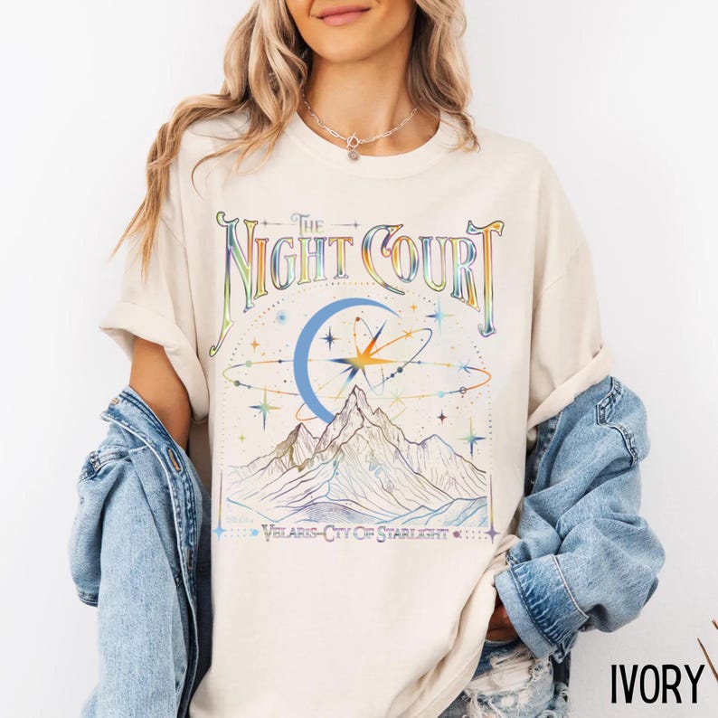 Op de afbeelding: Een cr&egrave;mekleurig T-shirt met een grafische print van een bergketen, een halve maan en sterren. De tekst "The Night Court" is gedrukt in een regenboogkleurige gradi&euml;nt. De tekst "Velaris - City of Starlight" is gedrukt onder de afbeelding. Het shirt is gemarkeerd met "Ivory".