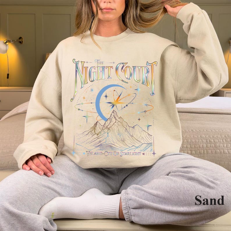 Op de afbeelding: Een beige sweatshirt met een grafische print van een bergketen, een halve maan en sterren. De tekst "The Night Court" is in regenboogkleuren boven de afbeelding gedrukt. De tekst "You are a City of Starlights..." is onder de afbeelding gedrukt.