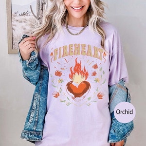 Puede incluir: Una camiseta de color lavanda con el texto "FIREHEART" en una fuente retro. Un coraz&oacute;n rojo estilizado con llamas est&aacute; centrado en la camiseta, rodeado de dise&ntilde;os florales. La camiseta est&aacute; siendo modelada por una persona que lleva una chaqueta vaquera.