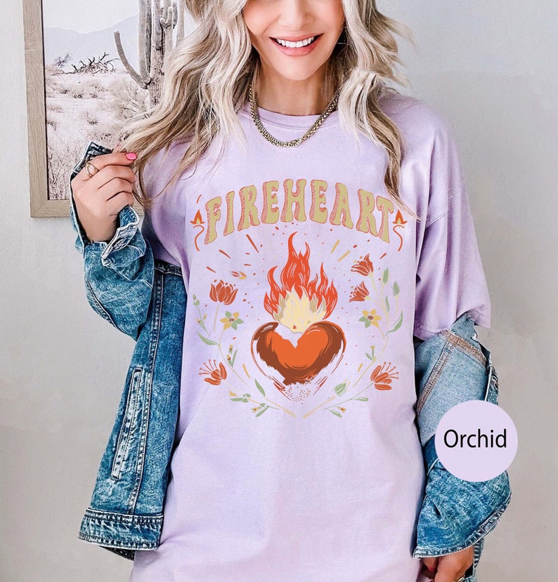 Puede incluir: Una camiseta de color lavanda con el texto "FIREHEART" en una fuente retro. Un coraz&oacute;n rojo estilizado con llamas est&aacute; centrado en la camiseta, rodeado de dise&ntilde;os florales. La camiseta est&aacute; siendo modelada por una persona que lleva una chaqueta vaquera.