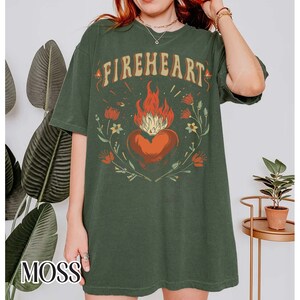 Puede incluir: Una camiseta verde de gran tama&ntilde;o con un estampado gr&aacute;fico de un coraz&oacute;n en llamas rodeado de flores. El texto "FIREHEART" est&aacute; impreso sobre el coraz&oacute;n.