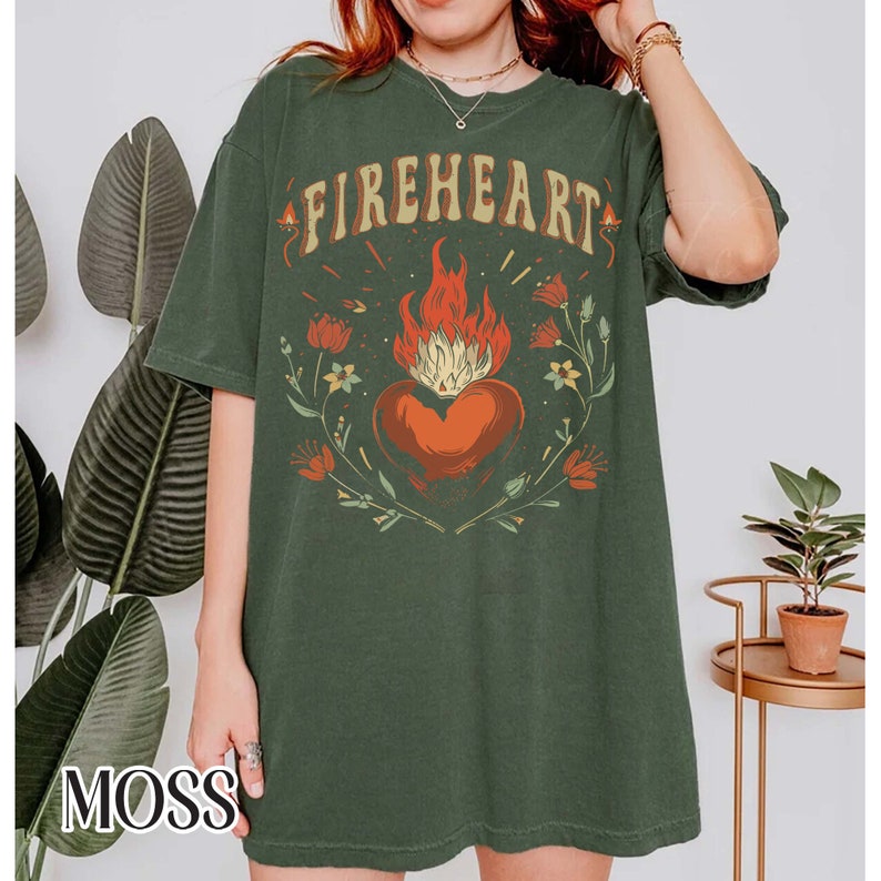 Puede incluir: Una camiseta verde de gran tama&ntilde;o con un estampado gr&aacute;fico de un coraz&oacute;n en llamas rodeado de flores. El texto "FIREHEART" est&aacute; impreso sobre el coraz&oacute;n.