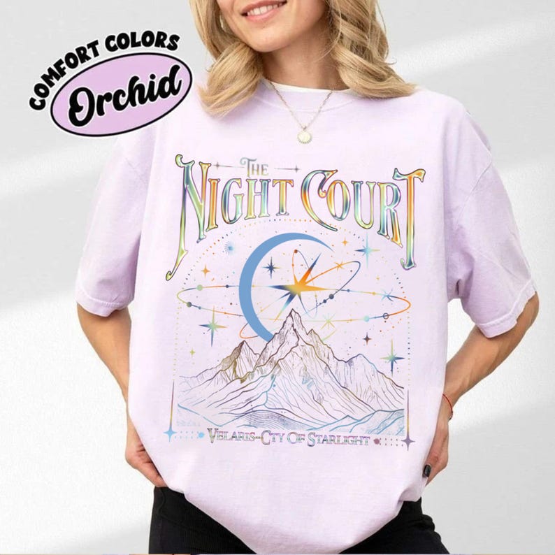 Op de afbeelding: Een lavendelkleurig T-shirt met een grafisch ontwerp van een bergketen, een halve maan en sterren. De tekst "The Night Court" is in regenboogkleuren boven de afbeelding gedrukt. De tekst "Velaris - City of Starlight" is onder de afbeelding gedrukt.