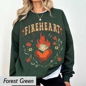Puede incluir: Sudadera verde bosque con un estampado gr&aacute;fico de un coraz&oacute;n en llamas rodeado de flores y el texto "FIREHEART".