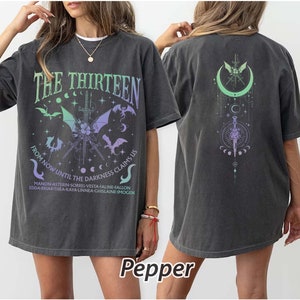 Peut inclure: Un t-shirt surdimensionné gris foncé avec un imprimé graphique d'une épée et de croissants de lune. Le texte "THE THIRTEEN" est imprimé en grande police au-dessus du graphique. Le texte "FROM NOW UNTIL THE DARKNESS CLAIMS US" est imprimé en police plus petite sous le graphique. Les noms "MANON-ASTERIN-SORREL-VESTA-FALINE-FALLON EDDA-BRIAR-THEA-KAYA-LINNEA-GHISLAINE-IMOGEN" sont imprimés en police encore plus petite sous le texte.