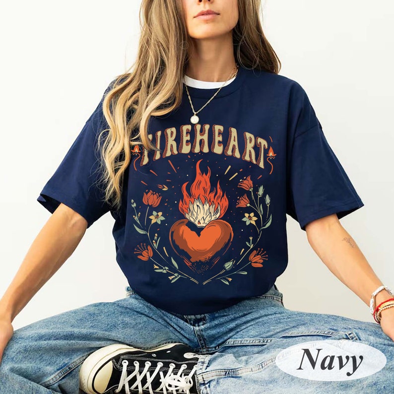Puede incluir: Una camiseta azul marino con un gr&aacute;fico de un coraz&oacute;n en llamas rodeado de flores. El texto "FIREHEART" est&aacute; por encima del coraz&oacute;n. La camiseta est&aacute; siendo modelada por una persona que lleva pantalones vaqueros azules.