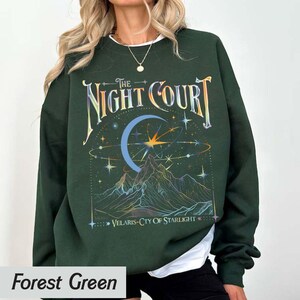 Op de afbeelding: Bosgroen sweatshirt met een grafische print van een halve maan, sterren en bergen. De tekst "The Night Court" en "Velaris-City of Starlight" staat op het sweatshirt gedrukt.