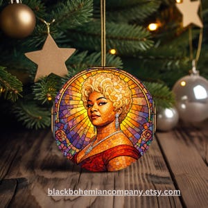 May include: Round Christmas ornament featuring a woman in a stained-glass style design. The ornament has a gold string for hanging. The woman has blonde hair, a red dress, and jewelry. The background is a mix of orange, purple, and yellow.