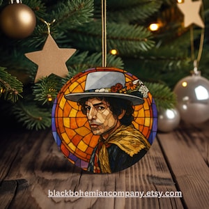 May include: Round Christmas ornament featuring a stained-glass style portrait of a man wearing a hat adorned with flowers. The ornament has a gold string for hanging and is set against a backdrop of a Christmas tree and wooden surface.