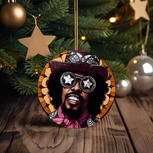 May include: Round Christmas ornament featuring a portrait of a smiling man with sunglasses, a hat, and a pink scarf. The ornament has a gold string for hanging and is set against a Christmas tree background.
