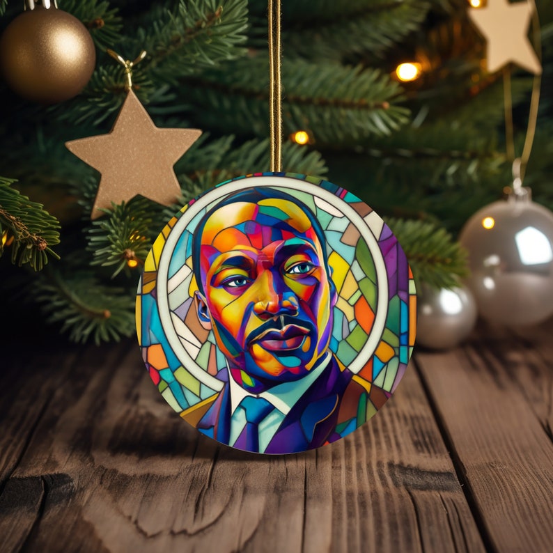 May include: A round Christmas ornament features a vibrant, stained-glass style portrait of Martin Luther King Jr. The artwork is predominantly composed of blues, yellows, oranges, and reds, creating a colorful and artistic depiction.  The ornament is perfect for celebrating the legacy of Dr. King during the holiday season. The image is set against a dark wooden background with a decorated Christmas tree visible in the soft focus background.