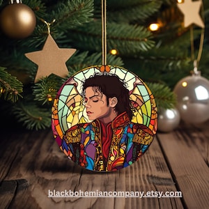 May include: A circular Christmas tree decoration with a stained-glass effect portrait of Michael Jackson. The ornament is suspended by a gold string. The background includes a Christmas tree and a wooden surface.