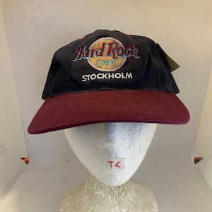 Op de afbeelding: Een zwarte honkbalpet met een bordeauxrode klep. De voorkant is voorzien van een geborduurd Hard Rock Cafe-logo in geel, rood en blauw, met het woord STOCKHOLM in witte letters onder het logo gestikt. De pet heeft een gestructureerde kroon en een platte klep.