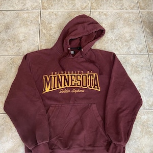 May include: A maroon hooded sweatshirt featuring long sleeves, a front kangaroo pocket, and a drawstring hood. The chest displays the text "UNIVERSITY OF MINNESOTA" in bold yellow letters with "Golden Gophers" written in a script font below it.