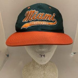 Könnte beinhalten: Eine dunkelgrüne Baseballkappe mit orangefarbenem Schirm. Auf der Vorderseite befindet sich ein in Orange und Weiß gestickter Schriftzug Miami Hurricanes in Schreibschrift. Die Kappe hat kleine orangefarbene Ösen an der Krone.