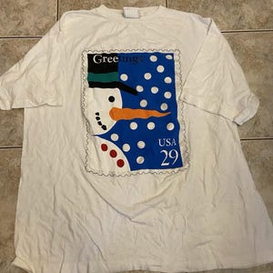 Puede incluir: Una camiseta blanca de manga corta con un gran estampado gráfico que representa un sello postal. El sello muestra un muñeco de nieve con sombrero negro y nariz de zanahoria naranja sobre un fondo azul con puntos blancos, con el texto Greetings, USA, 29.