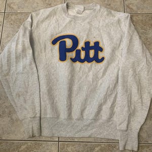 May include: A light grey heathered crewneck sweatshirt featuring the word Pitt in bold blue script lettering with a yellow outline across the chest. The garment has long sleeves, a ribbed collar, and ribbed cuffs, with a white tag at the neckline.