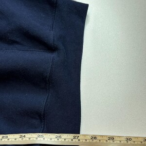 May include: A dark navy blue sweatshirt featuring a ribbed hemline. A yellow measuring tape is positioned horizontally across the bottom, showing a measurement extending past 30 inches, with the fabric edge aligned near the 27-inch mark.