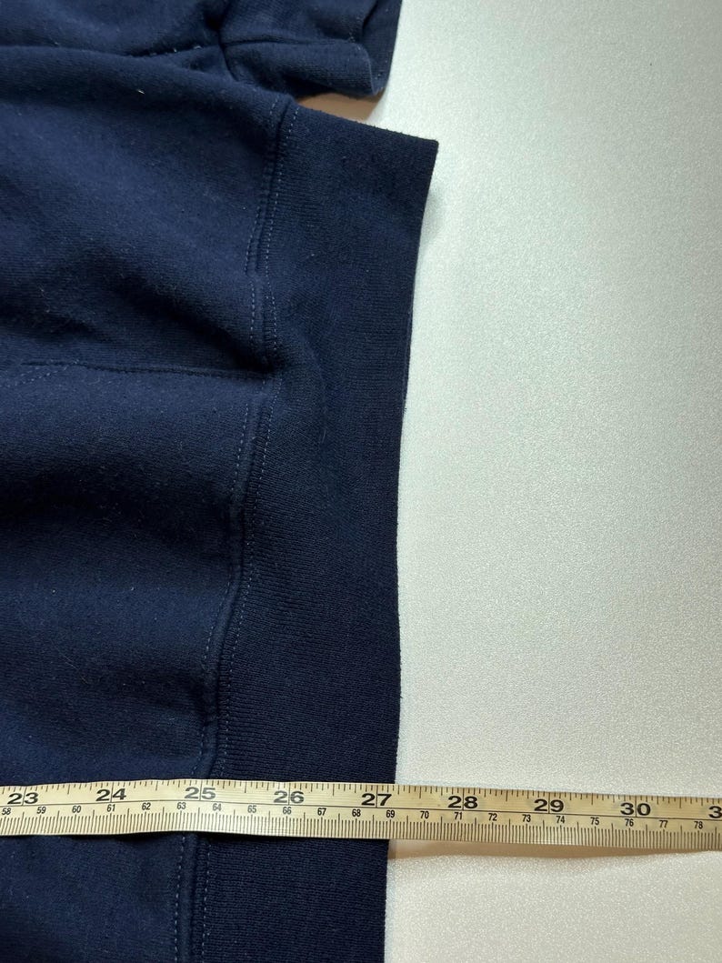 May include: A dark navy blue sweatshirt featuring a ribbed hemline. A yellow measuring tape is positioned horizontally across the bottom, showing a measurement extending past 30 inches, with the fabric edge aligned near the 27-inch mark.
