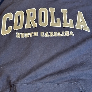 May include: A navy blue hooded sweatshirt featuring the word COROLLA in large, light gray block letters with a dark outline, and the text NORTH CAROLINA in smaller, matching light gray letters underneath. It includes a front kangaroo pocket.