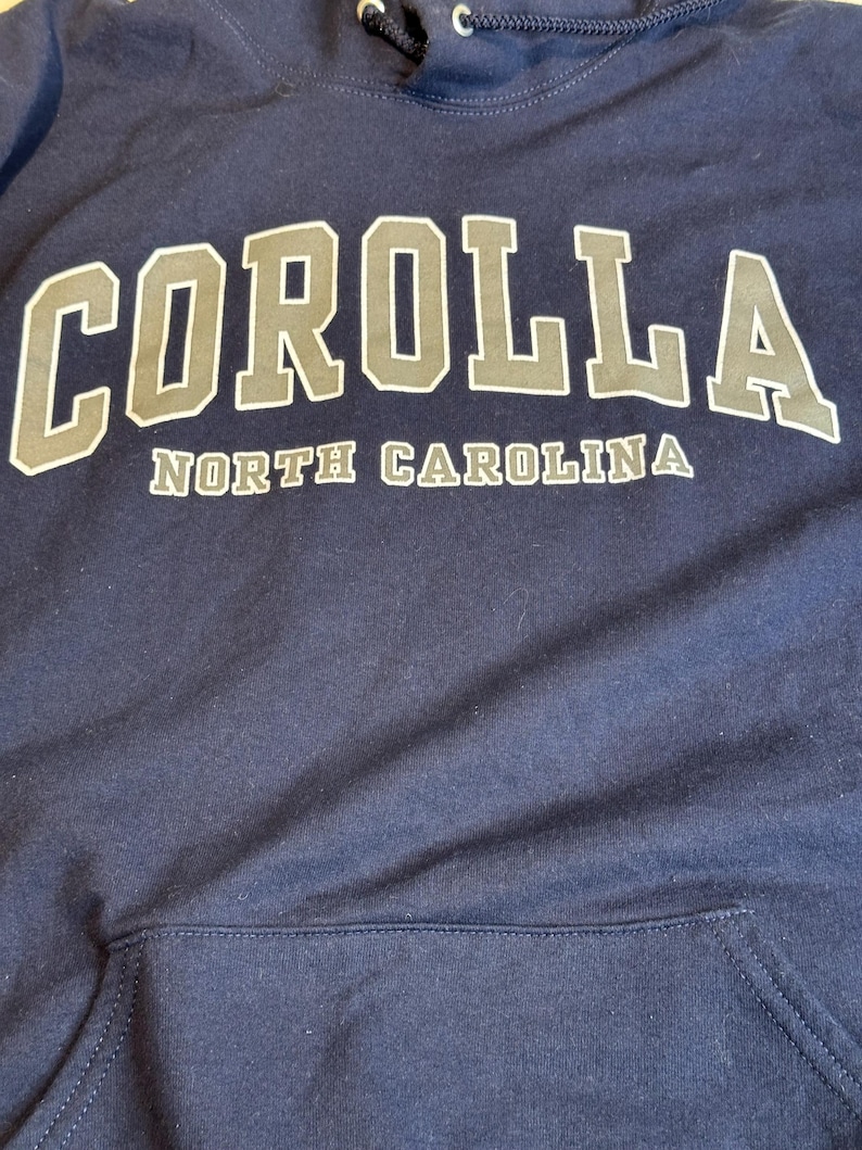 May include: A navy blue hooded sweatshirt featuring the word COROLLA in large, light gray block letters with a dark outline, and the text NORTH CAROLINA in smaller, matching light gray letters underneath. It includes a front kangaroo pocket.