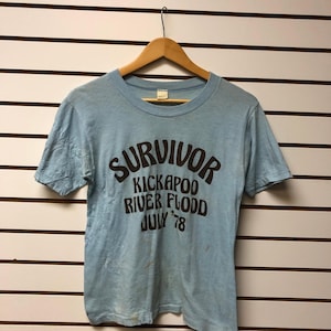 May include: A light blue short-sleeve t-shirt featuring dark brown text that reads SURVIVOR KICKAPOO RIVER FLOOD JULY '78. The shirt is displayed on a wooden hanger and has a crew neckline with a standard fit and visible signs of wear.