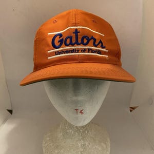 May include: An orange baseball cap featuring the word Gators in blue embroidery with white horizontal lines above and below the text. Below this, the text University of Florida is embroidered in blue. The cap has a structured crown and a stiff brim.