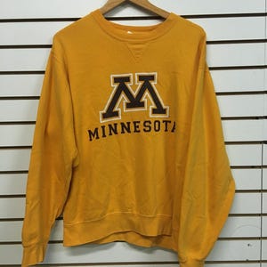 Puede incluir: Una sudadera de cuello redondo color dorado con un logotipo de letra M en bloque marrón y blanco sobre el texto MINNESOTA. El jersey de manga larga tiene un cuello acanalado con un detalle de costura triangular y puños y dobladillo acanalados.