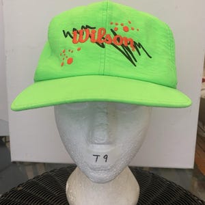 Könnte beinhalten: Eine neongrüne Baseballkappe mit dem in orangefarbenem Garn gestickten Wort Wilson. Das Design umfasst schwarze diagonale Pinselstrich-Akzente und kleine orangefarbene kreisförmige Punkte, die über dem Schirm auf der Vorderseite verteilt sind.