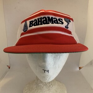 May include: A red and white trucker cap featuring horizontal stripes and the text Bahamas in dark blue lettering. The front panel includes embroidered icons of a sailboat and a palm tree. The cap has a solid red brim and white mesh side panels.