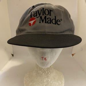 Könnte beinhalten: Eine graue Baseballkappe mit schwarzem Schirm und dem TaylorMade-Logo, das in schwarzer Schrift mit einem rot-weißen kreisförmigen Symbol auf der Vorderseite gestickt ist. Die Kappe hat eine strukturierte Krone mit einem kleinen schwarzen Ösendetail.